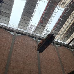 Usługi budowlane - Wnętrze budynku w trakcie rozbiórki: ceglana ściana, metalowa konstrukcja dachu z oknami, podnoszone elementy konstrukcyjne na haku. Widok z dołu.