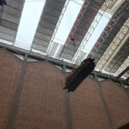 Wnętrze budynku w trakcie rozbiórki: ceglana ściana, metalowa konstrukcja dachu z oknami, podnoszone elementy konstrukcyjne na haku. Widok z dołu.