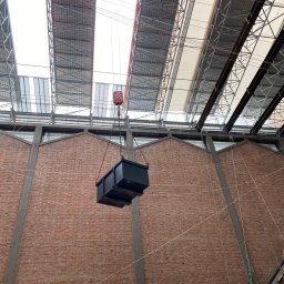 Usługi budowlane - Wnętrze budynku z ceglaną ścianą i rusztowaniami pod dachem. Czarny kontener podnoszony przez dźwig z czerwonym hakiem. Widoczne liny i elementy konstrukcyjne.