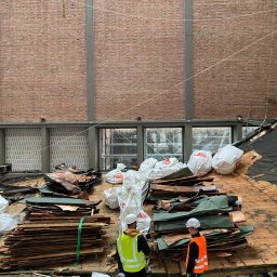 Usługi budowlane - Demontaż dachu z elementami miedzianymi. Dwóch robotników w odblaskowych kamizelkach i kaskach. Worki z odpadami i fragmenty konstrukcji. Ceglana ściana w tle.