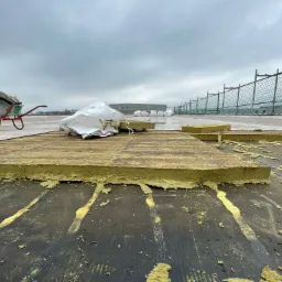 Ocieplanie dachu budynku przemysłowego: żółta wełna mineralna układana na czarnej membranie, w tle wózek i worki z materiałem, pochmurne niebo.