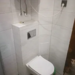 Wisząca miska WC z zabudowanym zbiornikiem, obłożona jasnymi, imitującymi drewno płytkami, z widocznymi przyłączami wodnymi i gniazdkiem elektrycznym w górnej części kadru.