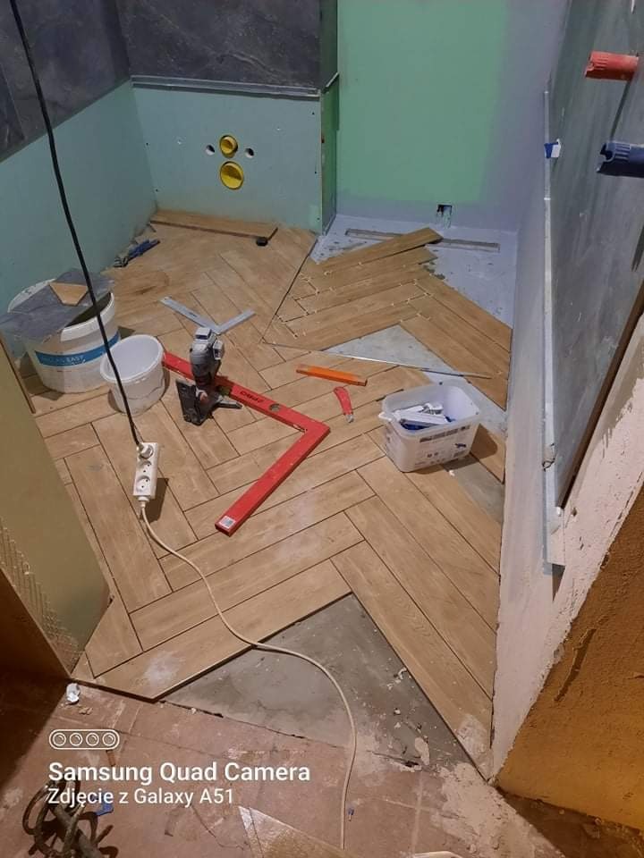 Układanie podłogi z płytek imitujących drewno w jodełkę w trakcie prac wykończeniowych w łazience, widoczne narzędzia glazurnicze, poziomica, puszki z klejem i gniazdko elektryczne.