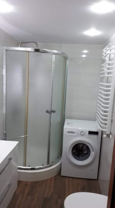 Wyremontowana łazienka z kabiną prysznicową narożną z matowego szkła, białą pralką i grzejnikiem drabinkowym na tle białych płytek i brązowej podłogi imitującej drewno.