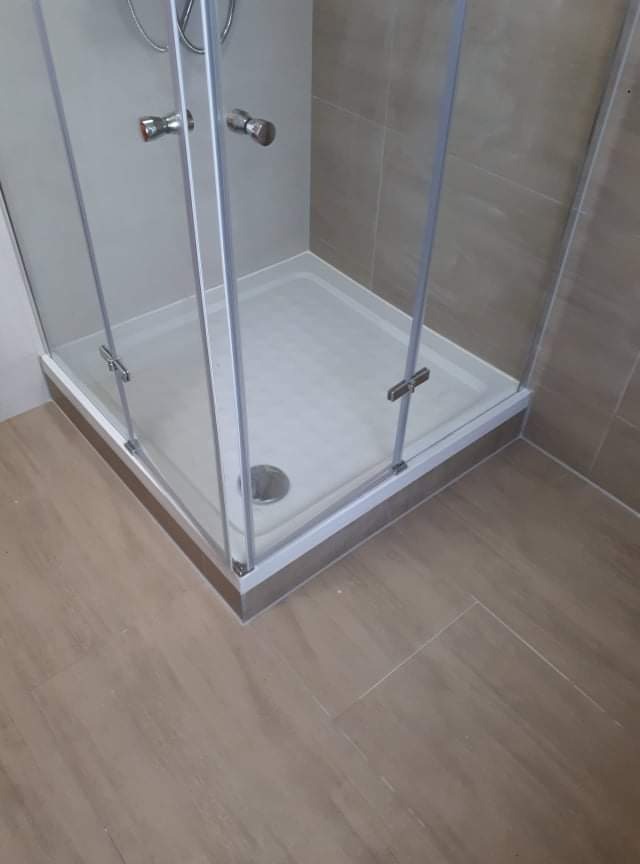 Narożna kabina prysznicowa z chromowanymi uchwytami i odpływem, uszczelniona silikonem, na podłodze płytki imitujące drewno, ściany wyłożone płytkami w odcieniach beżu.