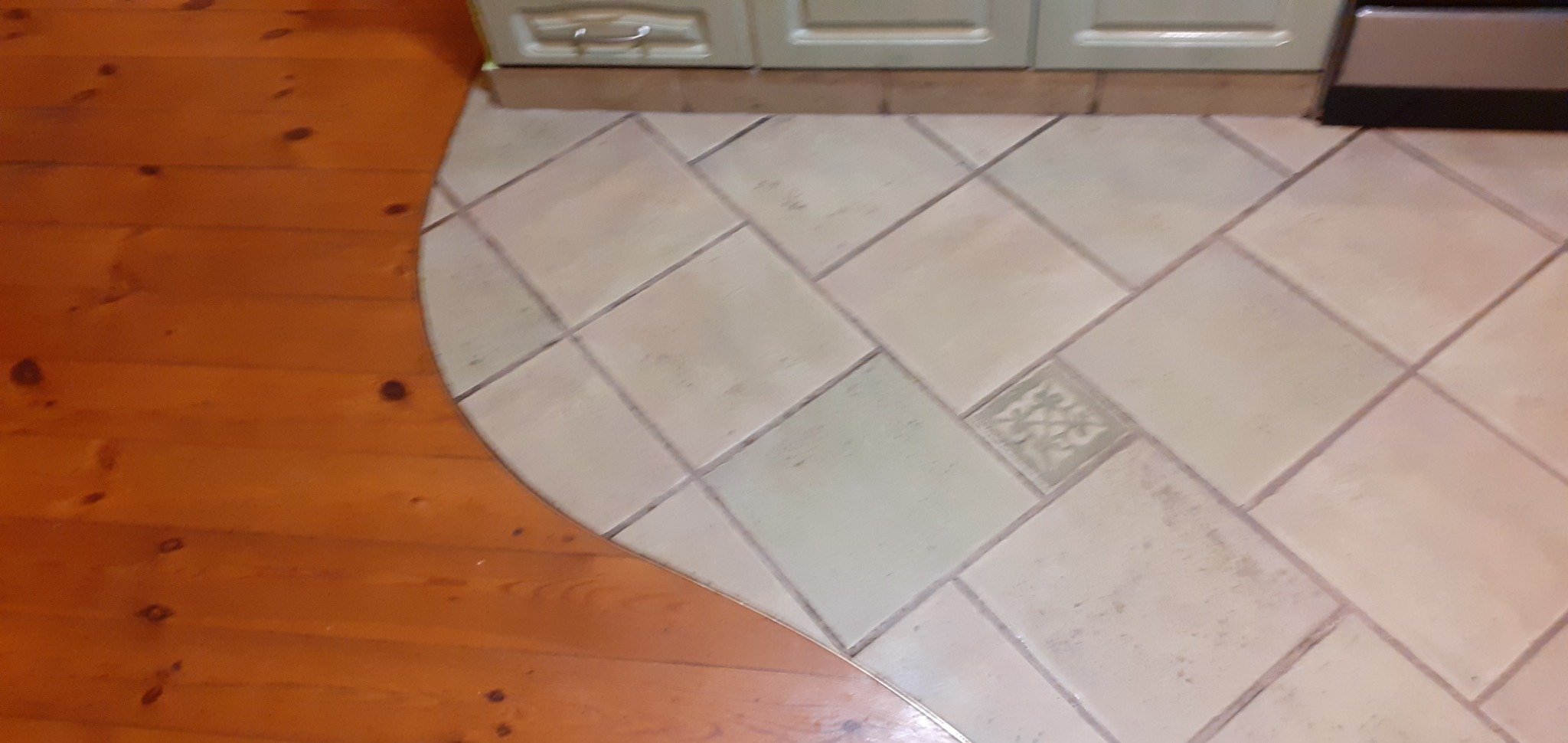 Połączenie drewnianej podłogi i ułożonych pod kątem jasnych, kwadratowych płytek w kuchni, z dekoracyjną płytką w centrum, widok z góry, fragment szafki w tle.