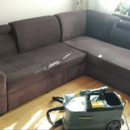 Narożnik z brązową tapicerką, częściowo wyczyszczony, obok odkurzacz piorący Karcher i wiadro, widoczne ślady czyszczenia na siedzisku.