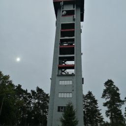 BACTECH Prace wysokościowe Marek Balcewicz - Wysoka wieża telekomunikacyjna z antenami i satelitami w Wałbrzychu, widziana z dołu na tle pochmurnego nieba, otoczona drzewami i ogrodzeniem.