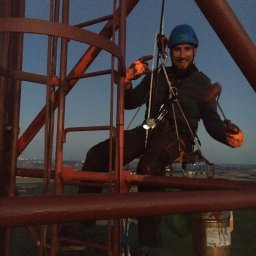 BACTECH Prace wysokościowe Marek Balcewicz - Uśmiechnięty pracownik w kasku i uprzęży, w trakcie prac wysokościowych na metalowej konstrukcji, z widocznym wiadrem z farbą i odległym miastem w tle, uchwycony przy zachodzącym słońcu.