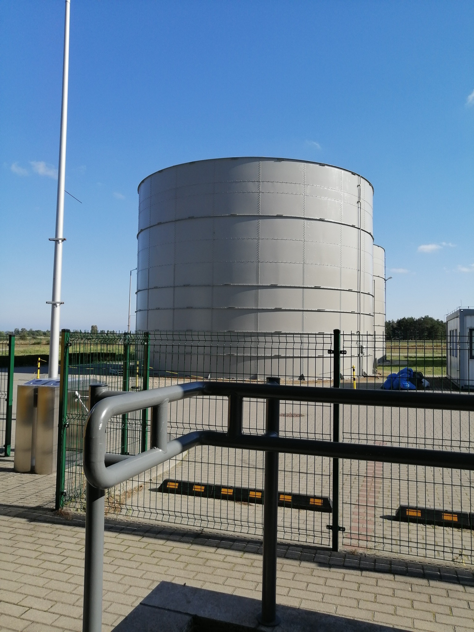 Dwa duże, stalowe zbiorniki przemysłowe na tle błękitnego nieba, widoczne zza metalowego ogrodzenia z elementami odblaskowymi.