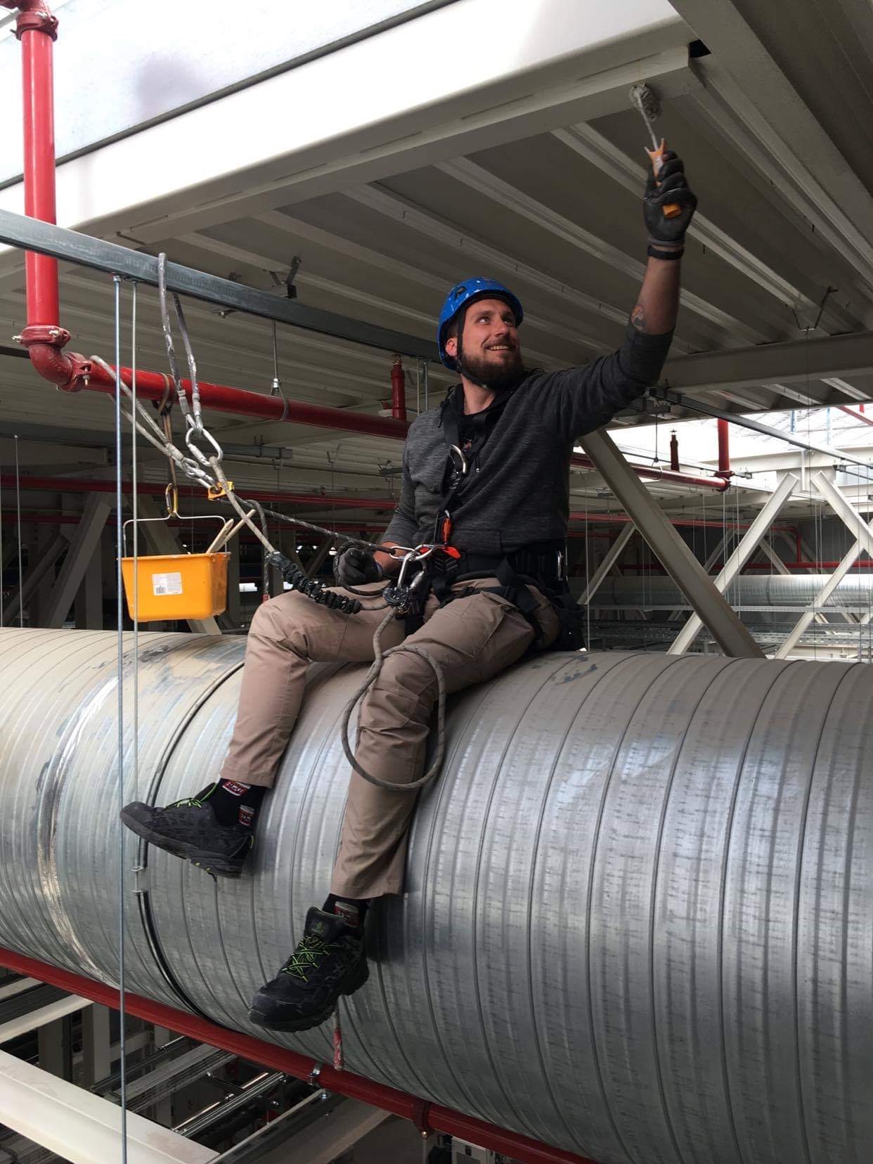 Mężczyzna w kasku i uprzęży, siedząc na metalowej rurze, maluje sufit pędzlem; widoczny żółty kubełek i czerwone rury pod sufitem.