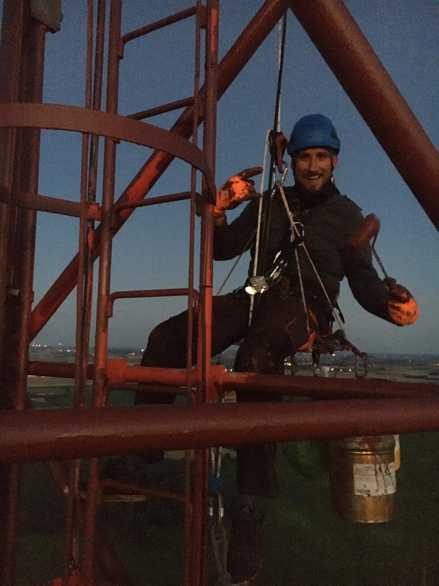 Uśmiechnięty pracownik w kasku i uprzęży, w trakcie prac wysokościowych na metalowej konstrukcji, z widocznym wiadrem z farbą i odległym miastem w tle, uchwycony przy zachodzącym słońcu.