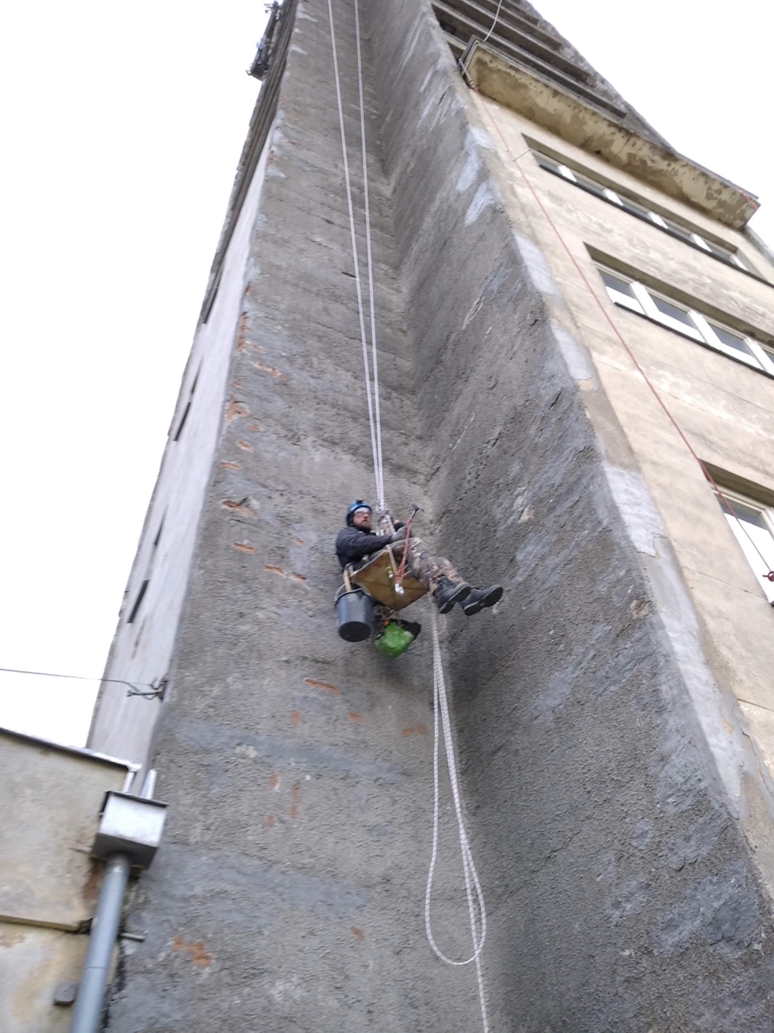 Pracownik w uprzęży, wiszący na linach, na tle wysokiego, szarego budynku z widocznymi ubytkami tynku i kilkoma oknami, trzymający wiadro i narzędzia.