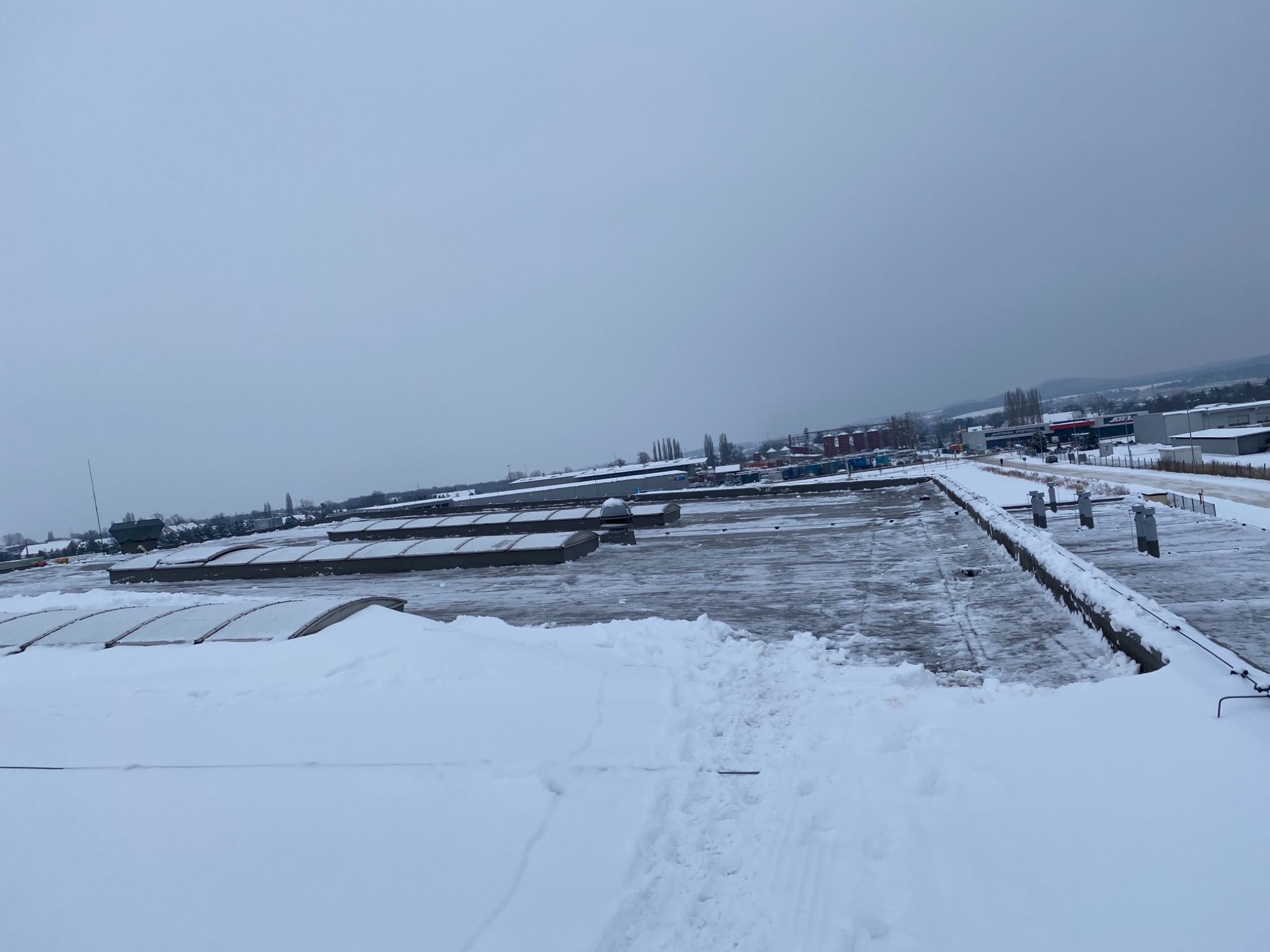 Śnieżny dach przemysłowy z widocznymi świetlikami i śladami stóp, pochmurne niebo w tle.