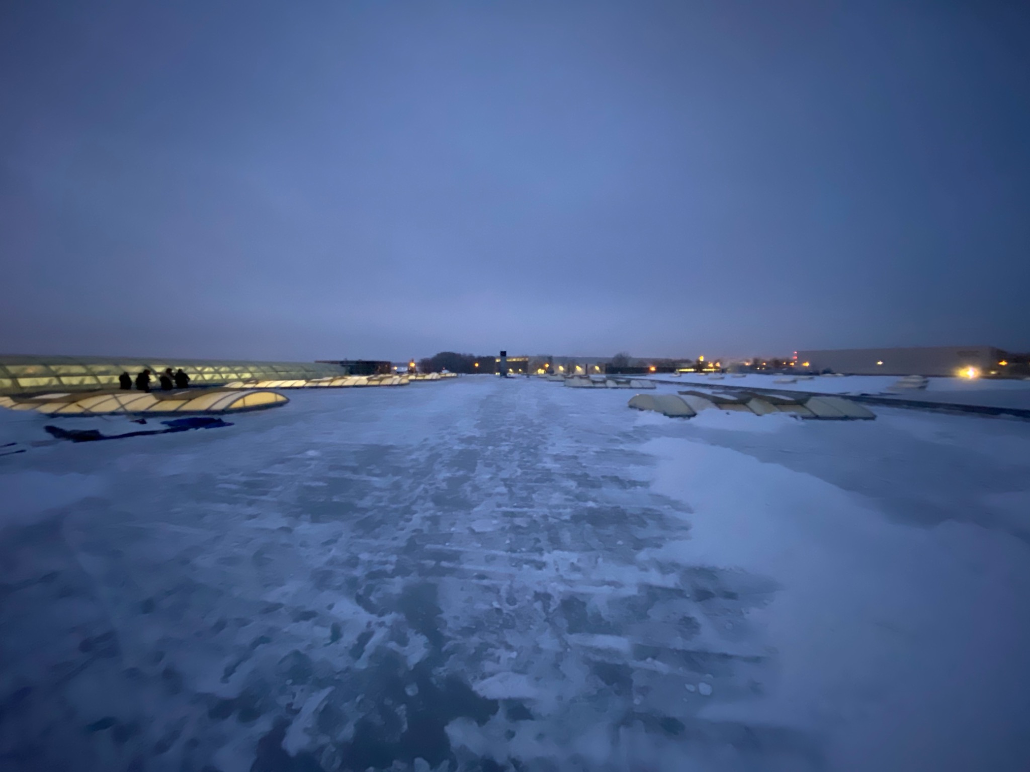 Rozległy, lekko ośnieżony dach z widocznymi świetlikami i sylwetkami osób na jednym z nich, sfotografowany o zmierzchu.