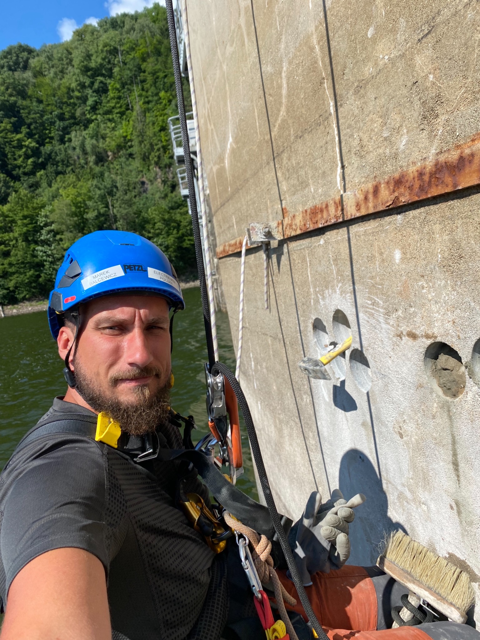 Pracownik w uprzęży alpinistycznej, w kasku Petzl, czyści betonową ścianę zapory, używając pędzla, widoczne otwory w ścianie i zardzewiałe elementy metalowe, w tle zalesione wzgórze i woda.