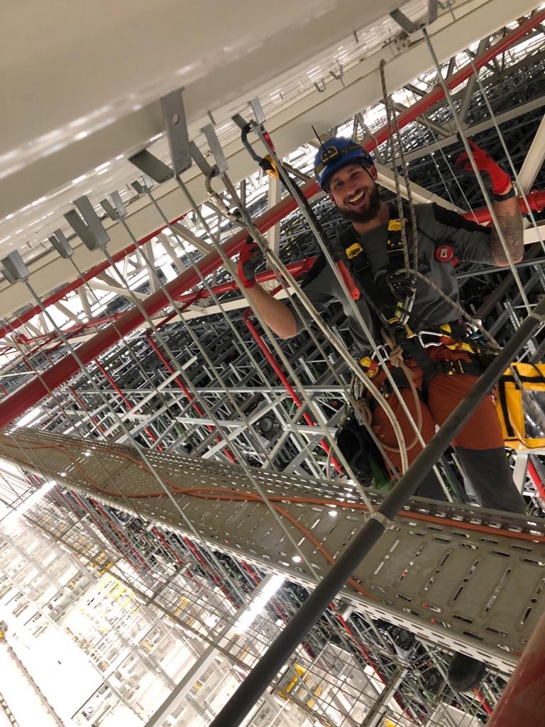 Uśmiechnięty pracownik w kasku i uprzęży, z uniesioną dłonią w czerwonej rękawicy, na tle skomplikowanej konstrukcji stalowej z rurami i kablami, widziany z dołu.