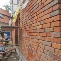 Wojciech Chałubowicz - Świeżo wymurowana ściana z czerwonej cegły, widoczne rusztowanie i pracownik w tle, budynek w stanie surowym otwartym.