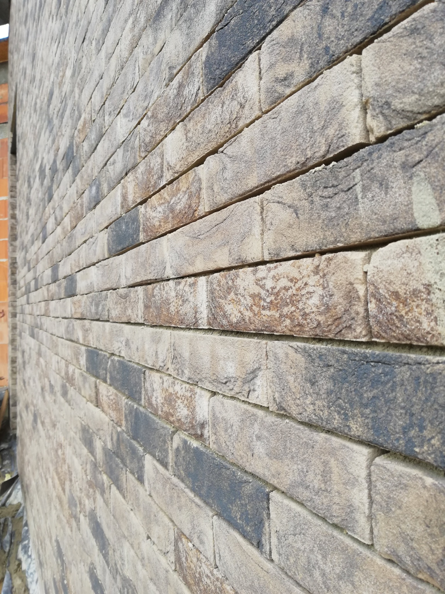 Ściana zewnętrzna z dekoracyjnej cegły klinkierowej w odcieniach szarości i beżu, ułożona poziomo z widoczną zaprawą, z fragmentem surowej ściany z czerwonej cegły w tle.