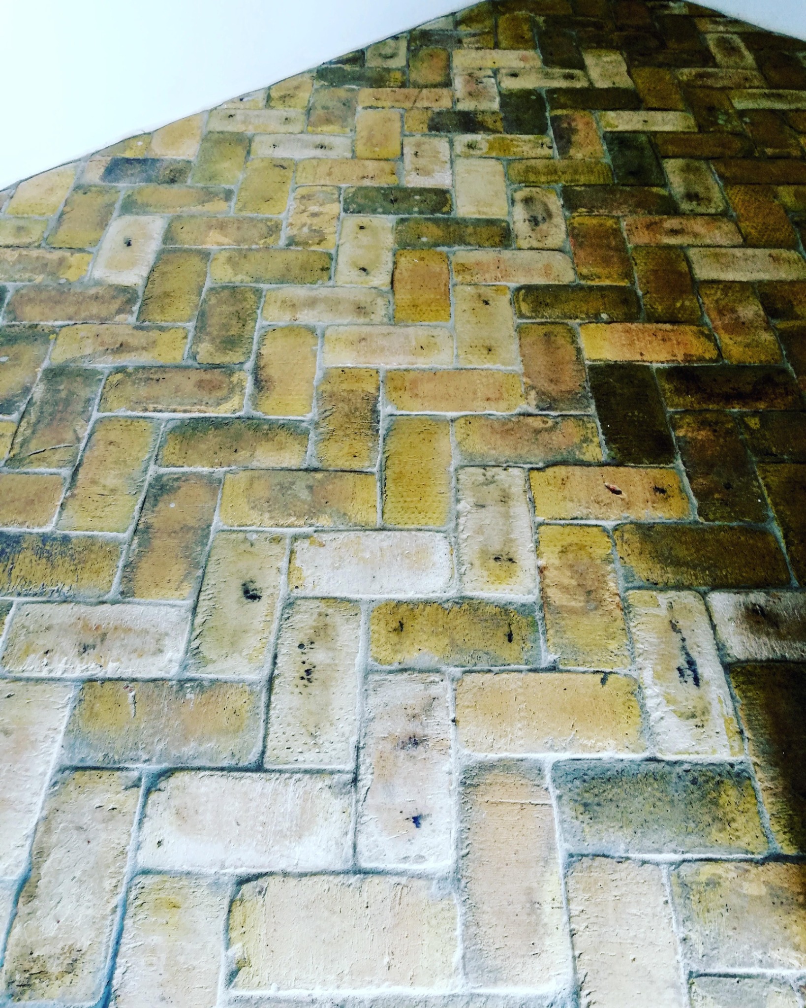 Murowana ściana z nieregularnych, żółto-beżowych cegieł ułożonych w dekoracyjny wzór, widoczna pod kątem od dołu, z fragmentem białego sufitu w górnym rogu.