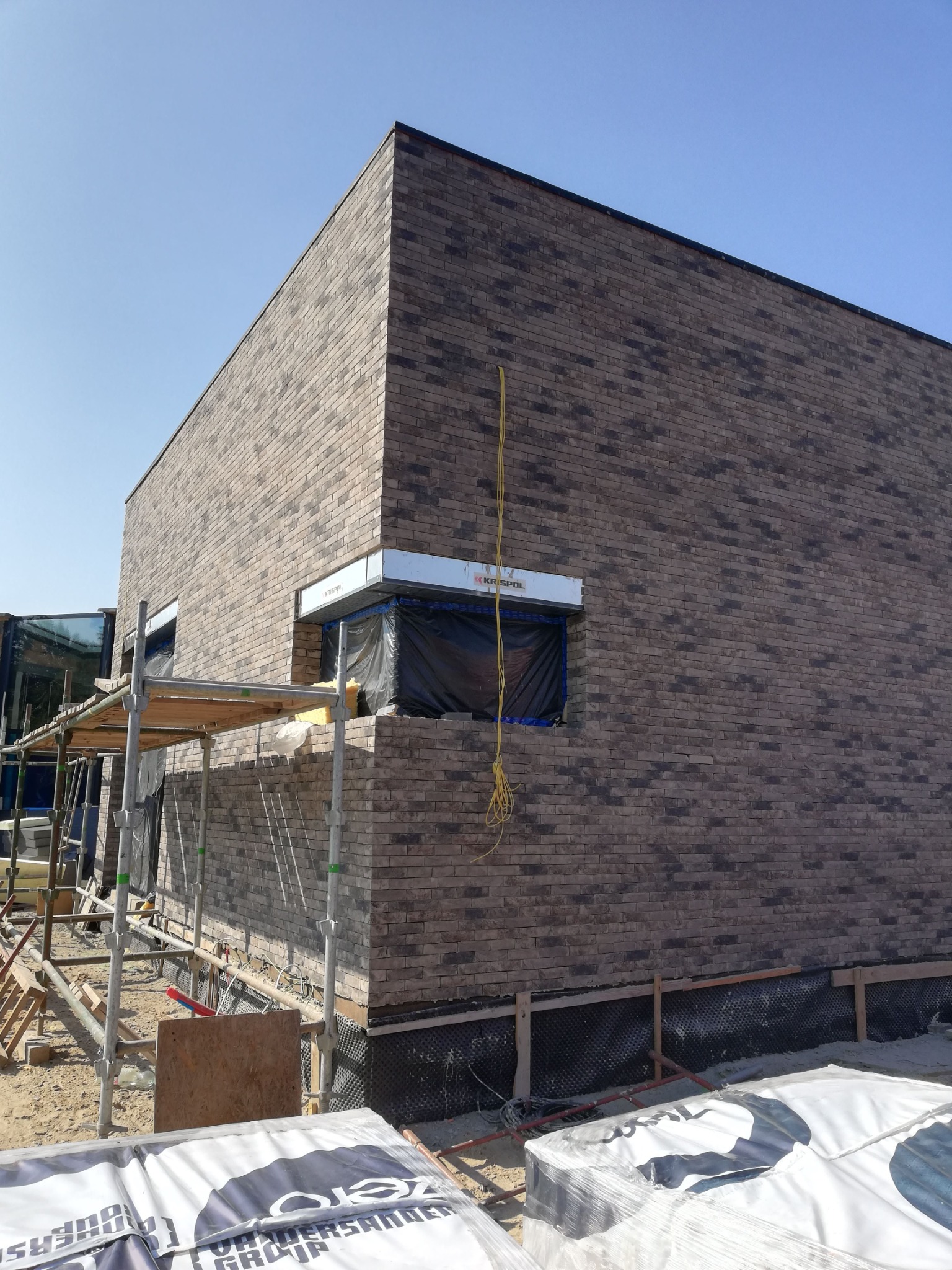 Wznoszona ściana z brązowej cegły klinkierowej na nowo budowanym domu, widoczne rusztowania i zabezpieczone okno folią, materiały budowlane na pierwszym planie.