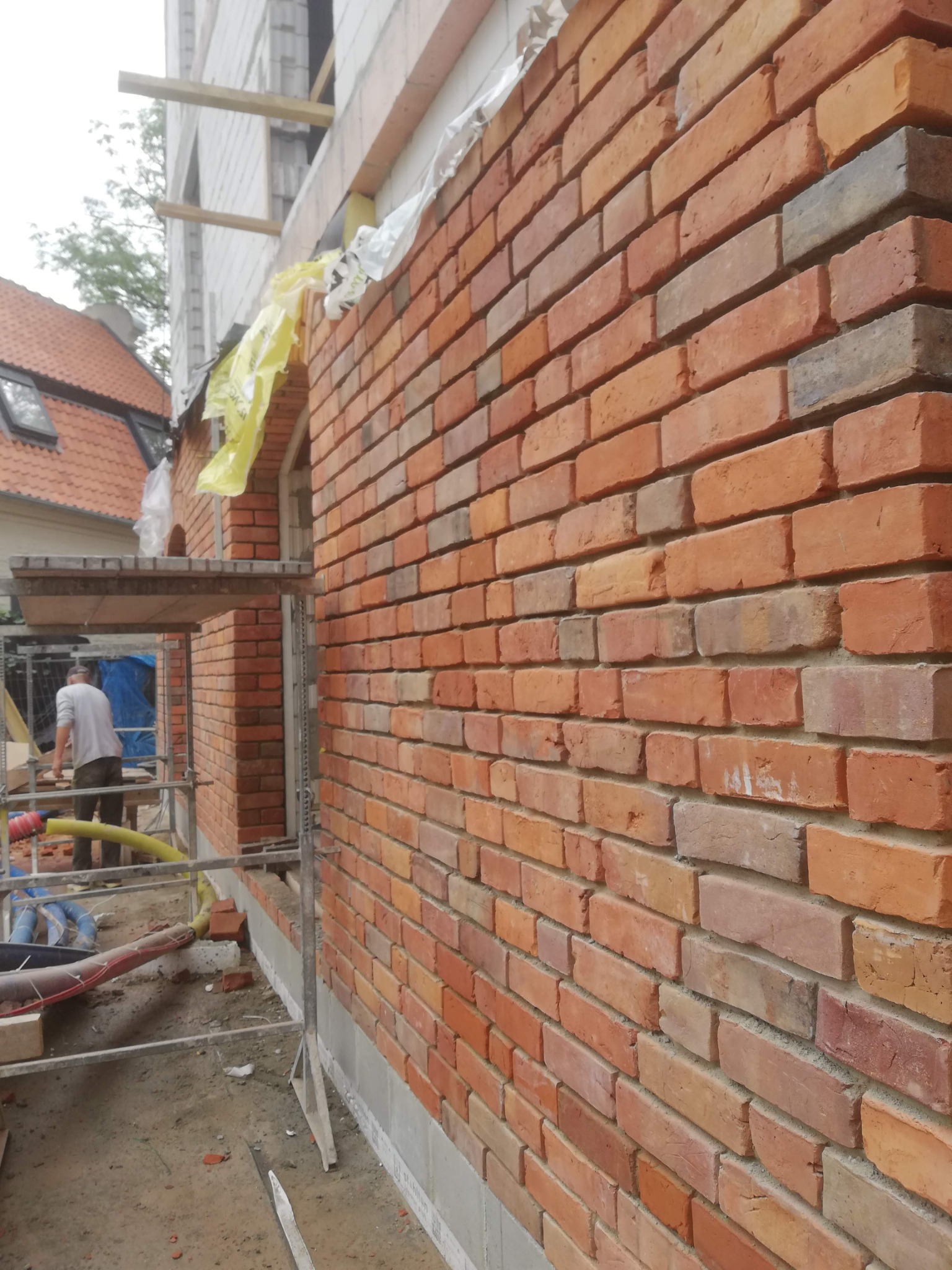 Świeżo wymurowana ściana z czerwonej cegły, widoczne rusztowanie i pracownik w tle, budynek w stanie surowym otwartym.