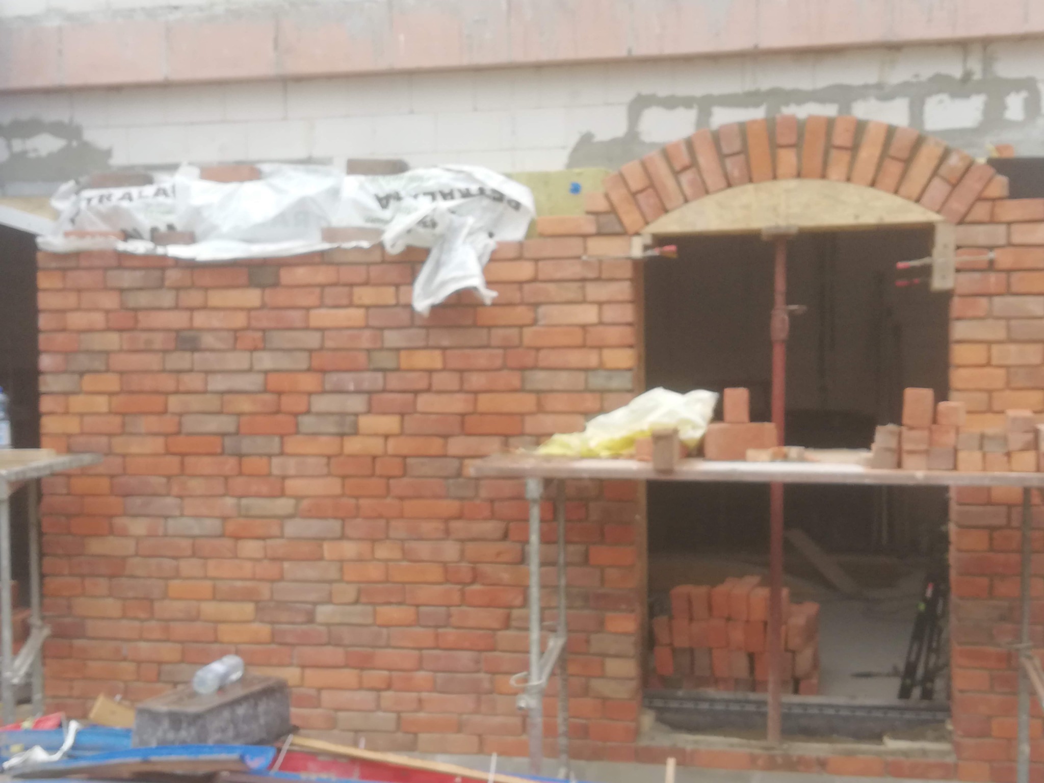 Budowa ściany z czerwonej cegły z łukowym otworem okiennym, widoczne rusztowanie i materiały budowlane.