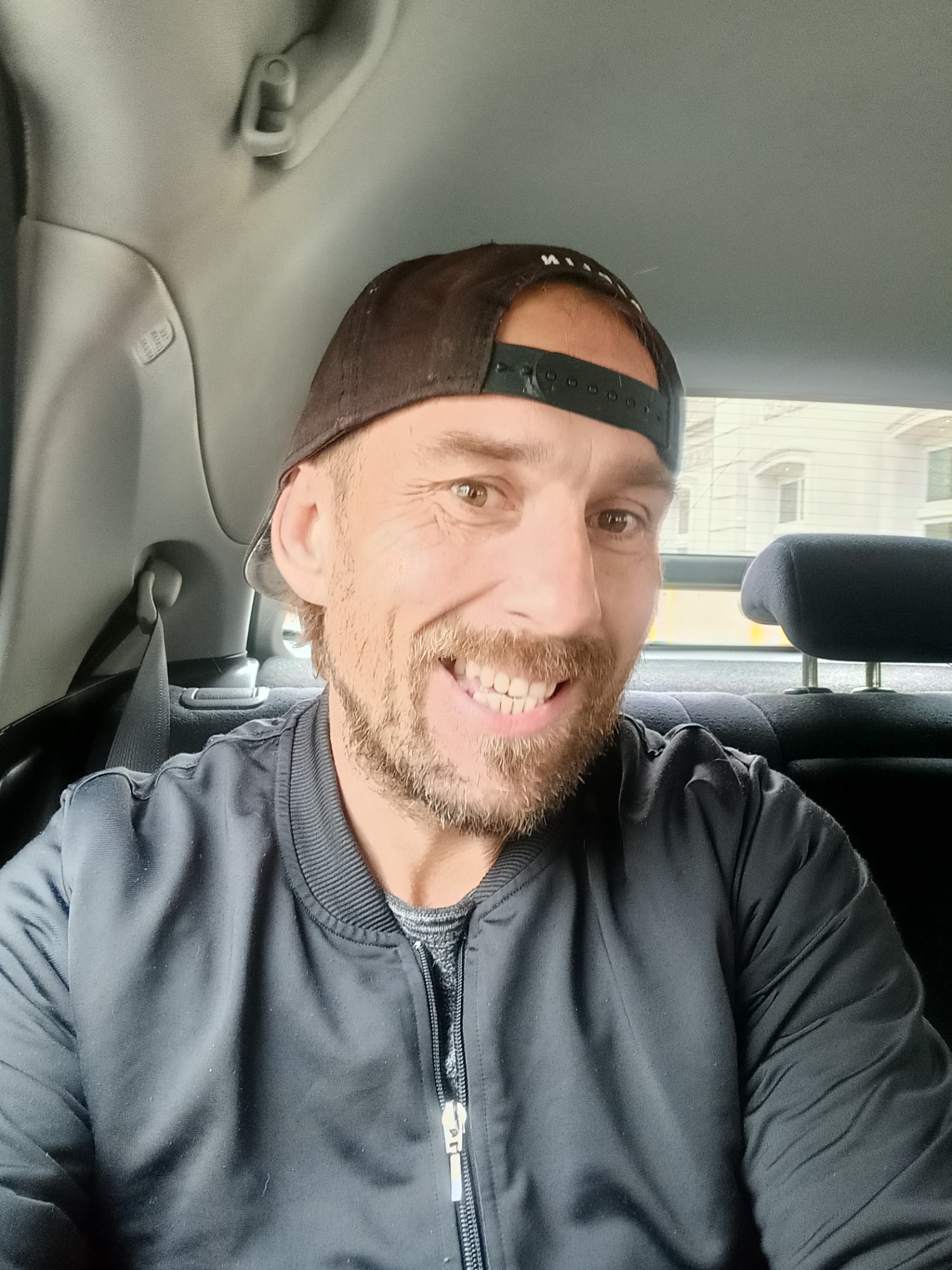 Uśmiechnięty mężczyzna w czarnej kurtce i czapce z daszkiem, siedzący na tylnym siedzeniu samochodu z widokiem na budynek przez okno.