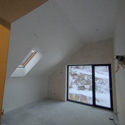 NUMERNABIS PROJEKTY SPÓŁKA Z OGRANICZONĄ ODPOWIEDZIALNOŚCIĄ - Surowe wnętrze poddasza w Krakowie z oknami dachowymi i dużym oknem z widokiem na zimowy krajobraz, ściany przygotowane pod malowanie, widoczne ślady po szpachlowaniu.