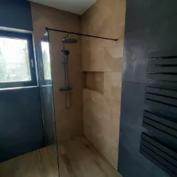 Nowoczesna kabina prysznicowa z geometrycznymi płytkami w odcieniach beżu, czarną armaturą i grzejnikiem drabinkowym, oświetlona światłem dziennym wpadającym przez okno.