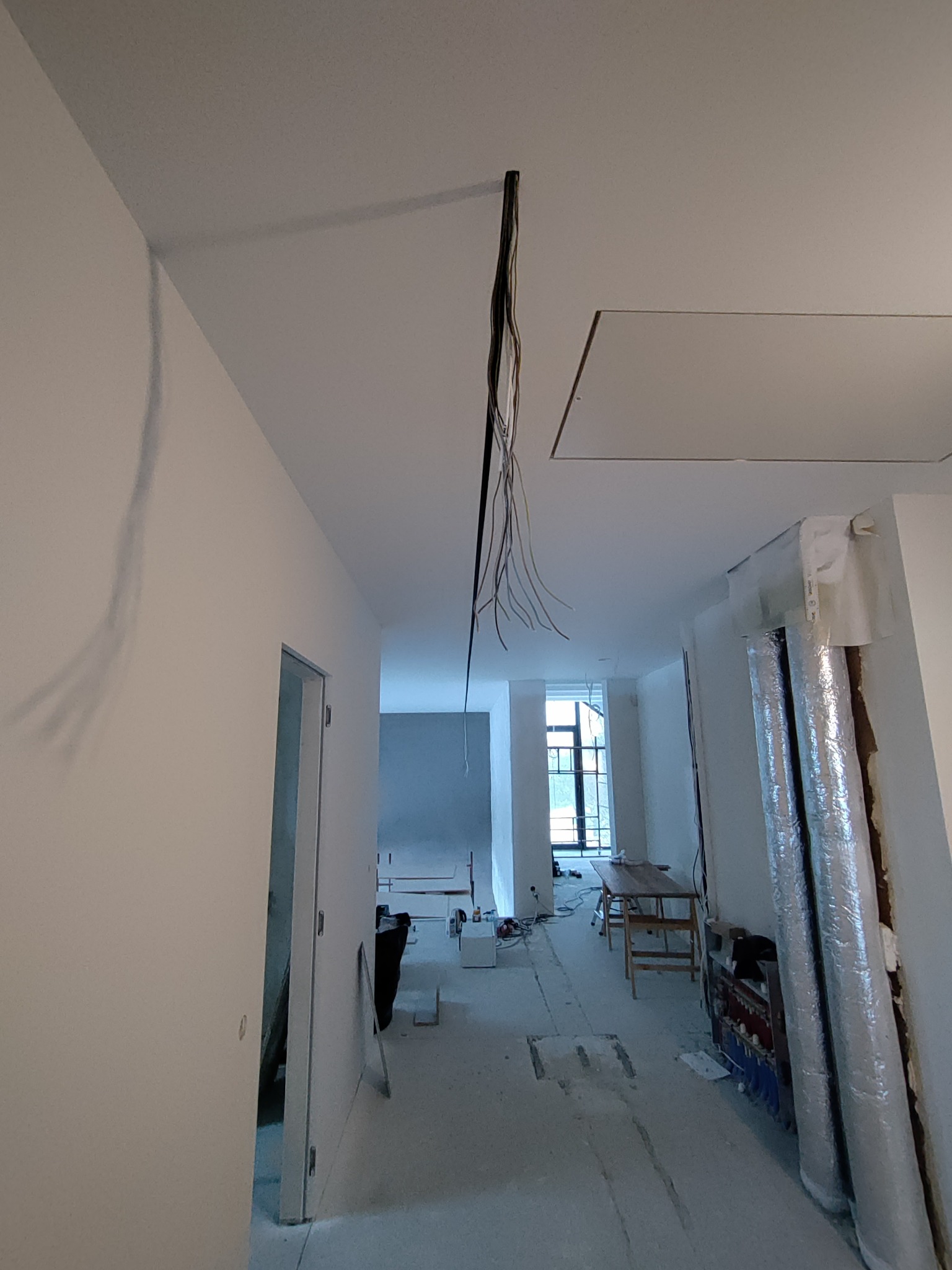 Wnętrze w trakcie remontu: widoczne przewody elektryczne zwisające z sufitu, ślady po gładzi na ścianie, rury w izolacji oraz przygotowane podłoże pod posadzkę.