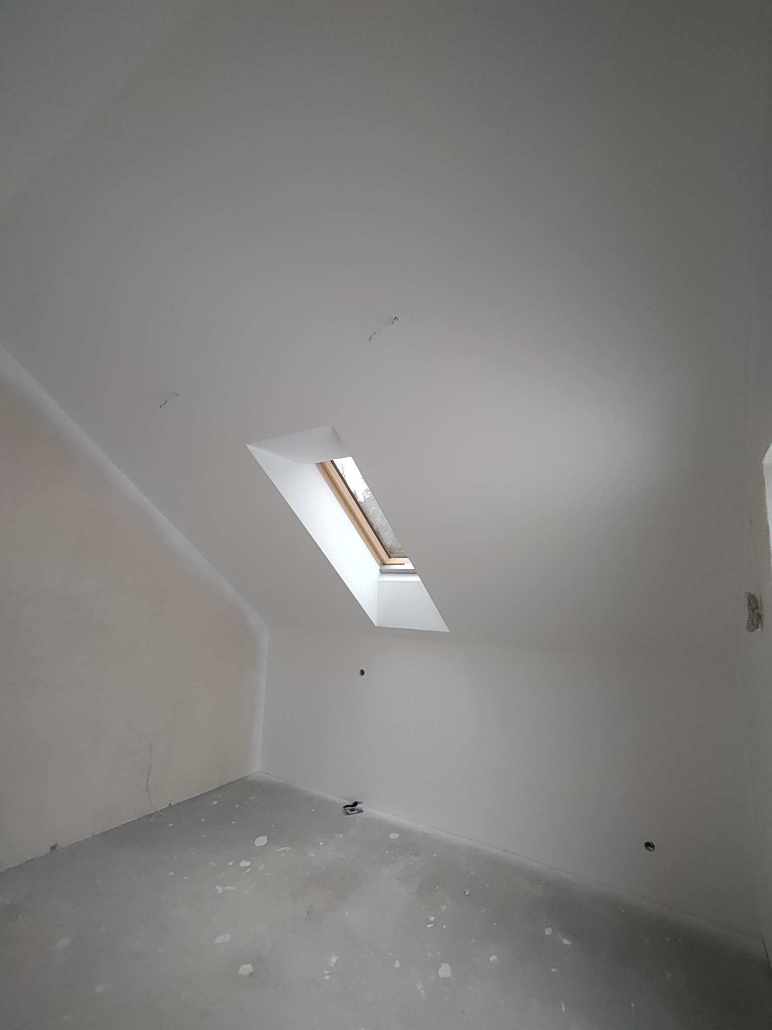 Poddasze z oknem dachowym po nałożeniu gładzi, widoczne niedoskonałości na podłodze i ścianach przed dalszym wykończeniem.