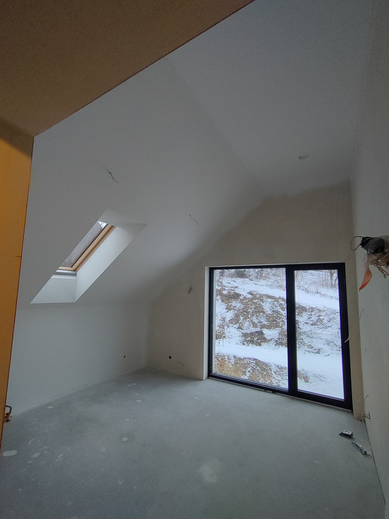 Surowe wnętrze poddasza w Krakowie z oknami dachowymi i dużym oknem z widokiem na zimowy krajobraz, ściany przygotowane pod malowanie, widoczne ślady po szpachlowaniu.