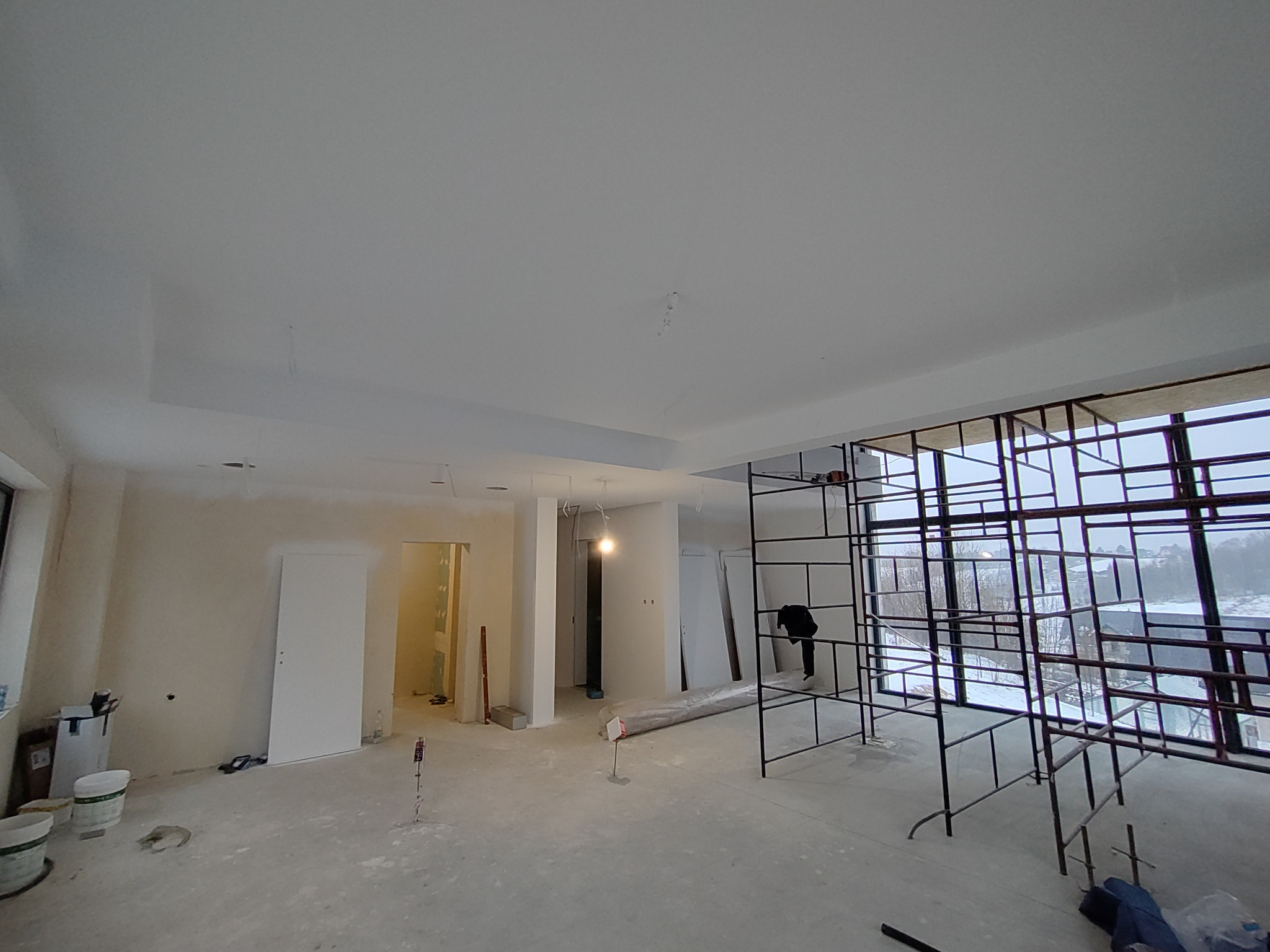 Przestronne wnętrze w trakcie prac wykończeniowych, widoczne rusztowania przy oknach, surowy beton na podłodze i częściowo wykończone ściany, zarys sufitu podwieszanego.