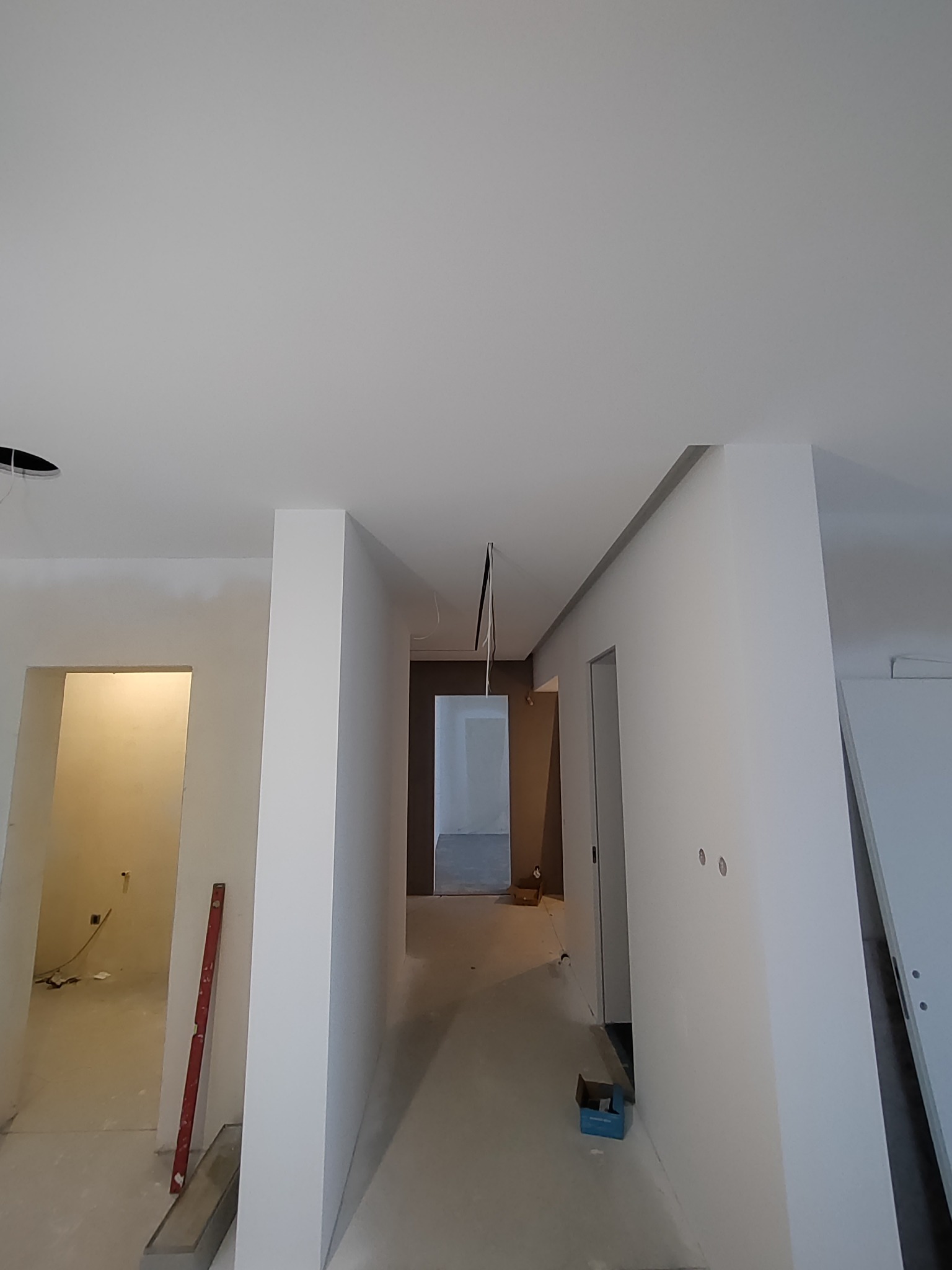 Korytarz w trakcie prac wykończeniowych, z widocznymi gładkimi, białymi ścianami, poziomica oparta o ścianę, surowe oświetlenie i otwarte przejścia do innych pomieszczeń.
