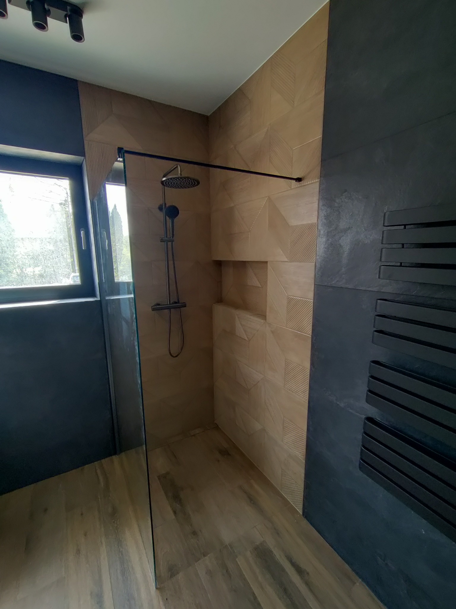 Nowoczesna kabina prysznicowa z geometrycznymi płytkami w odcieniach beżu, czarną armaturą i grzejnikiem drabinkowym, oświetlona światłem dziennym wpadającym przez okno.
