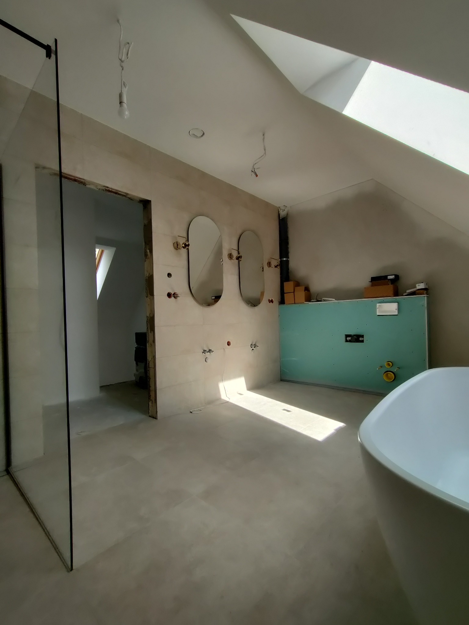 Łazienka na poddaszu w trakcie wykańczania: widoczne płytki na ścianach, dwie owalne lustra z elementami w kolorze złota, kabina prysznicowa ze szkła, fragment wanny oraz przygotowana ściana...