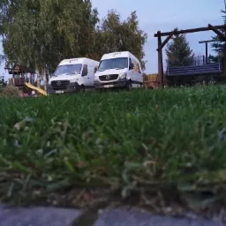 Dwa białe busy GLS zaparkowane na trawie przed domem z placem zabaw i huśtawką.