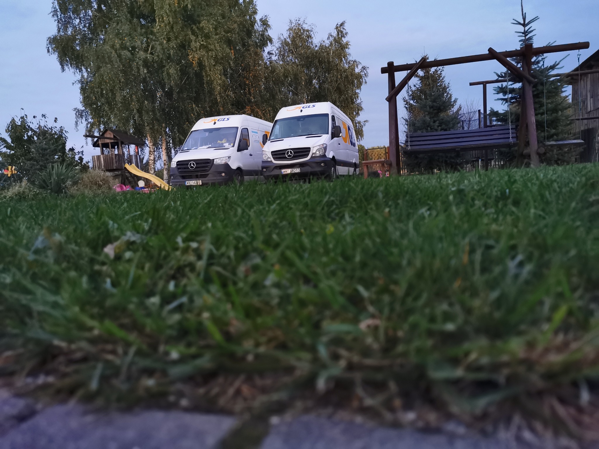 Dwa białe busy GLS zaparkowane na trawie przed domem z placem zabaw i huśtawką.