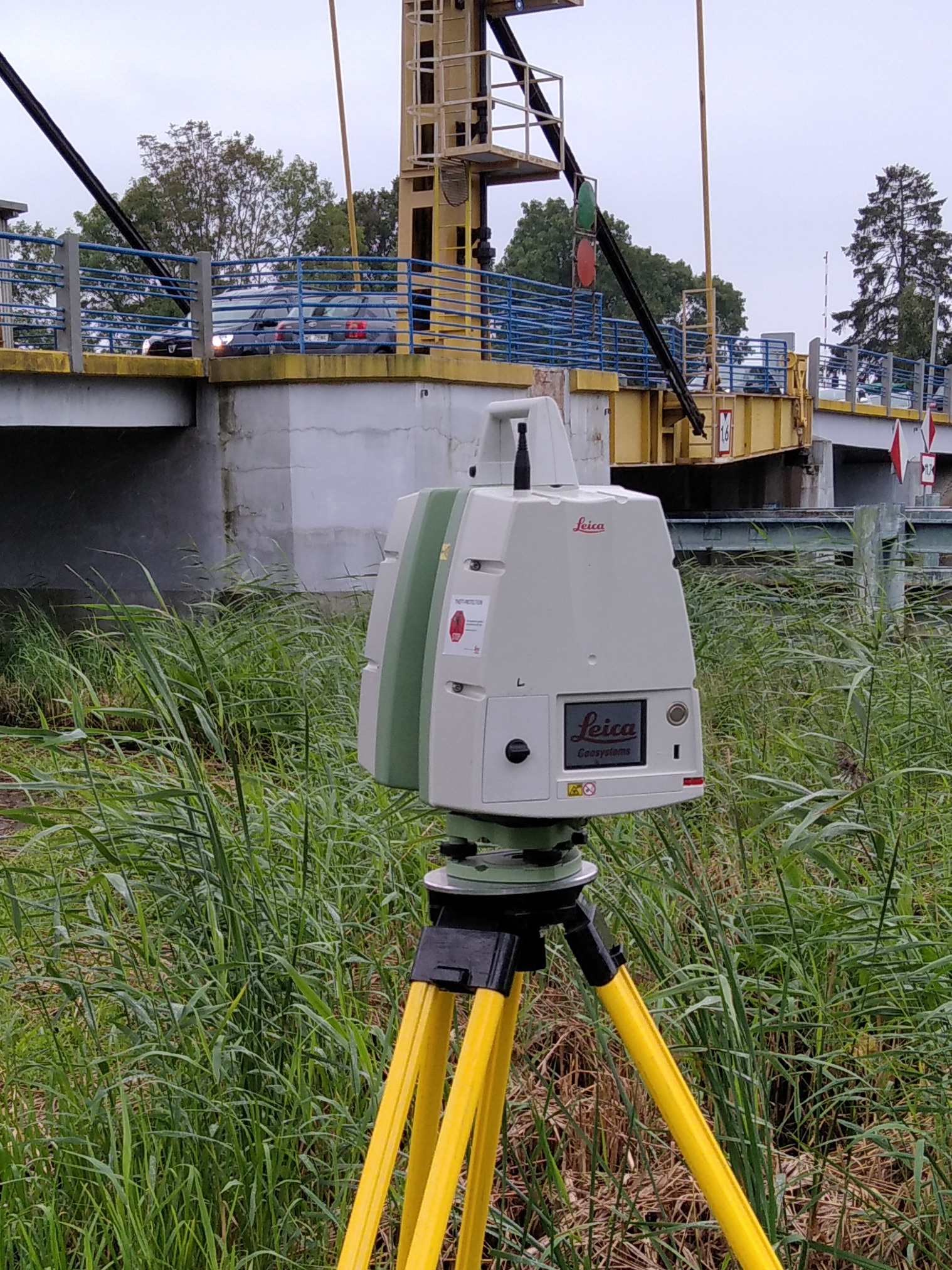 Skaner laserowy Leica na żółtym statywie, ustawiony w wysokiej trawie, z widokiem na most zwodzony i przejeżdżający samochód.