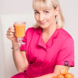 Uśmiechnięta blondynka w różowym uniformie trzyma szklankę pomarańczowego soku, obok talerz z mandarynkami na białym stole.