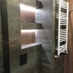 Nowoczesna łazienka z ciemnymi, teksturalnymi płytkami, podświetlanymi półkami wnękowymi i białym grzejnikiem drabinkowym.