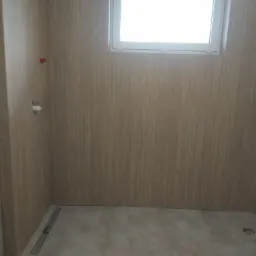 Wykończona łazienka z drewnopodobnymi panelami ściennymi, szarą podłogą z odpływem liniowym i oknem w białej ramie, widoczne przyłącza wodne.