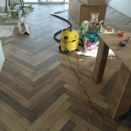 Ułożona podłoga z paneli w jodełkę, widoczny odkurzacz przemysłowy, kartony, narzędzia i materiały budowlane, okno balkonowe w tle.