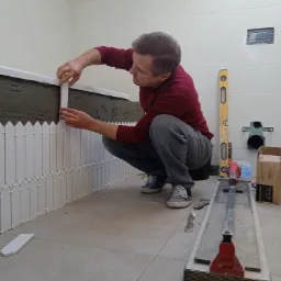 Glazurnik w trakcie układania białych, pionowych płytek imitujących płotek wokół wanny w łazience, z użyciem poziomicy i gilotyny do płytek.