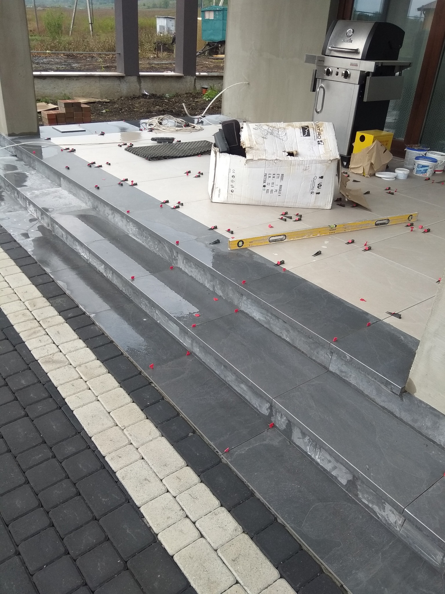 Kładzenie ciemnoszarych płytek na schodach wejściowych, widoczne poziomica, krzyżyki dystansowe i karton po płytkach, obok kostka brukowa w biało-czarne pasy, w tle grill i elementy ogrodu.
