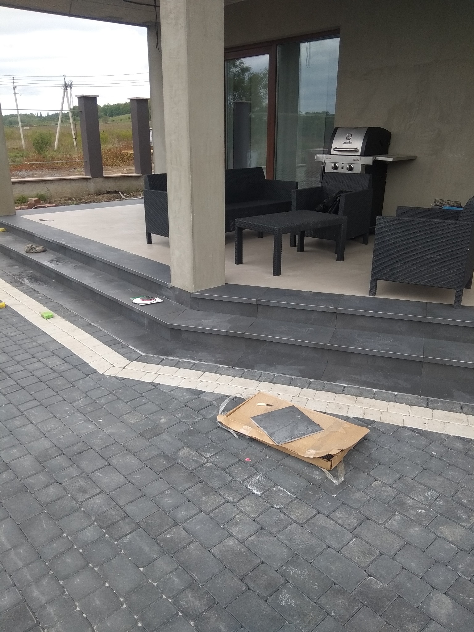 Zewnętrzny taras z ciemnoszarą kostką brukową i schodami wyłożonymi płytkami, widoczne meble ogrodowe i grill, karton z płytkami na pierwszym planie.