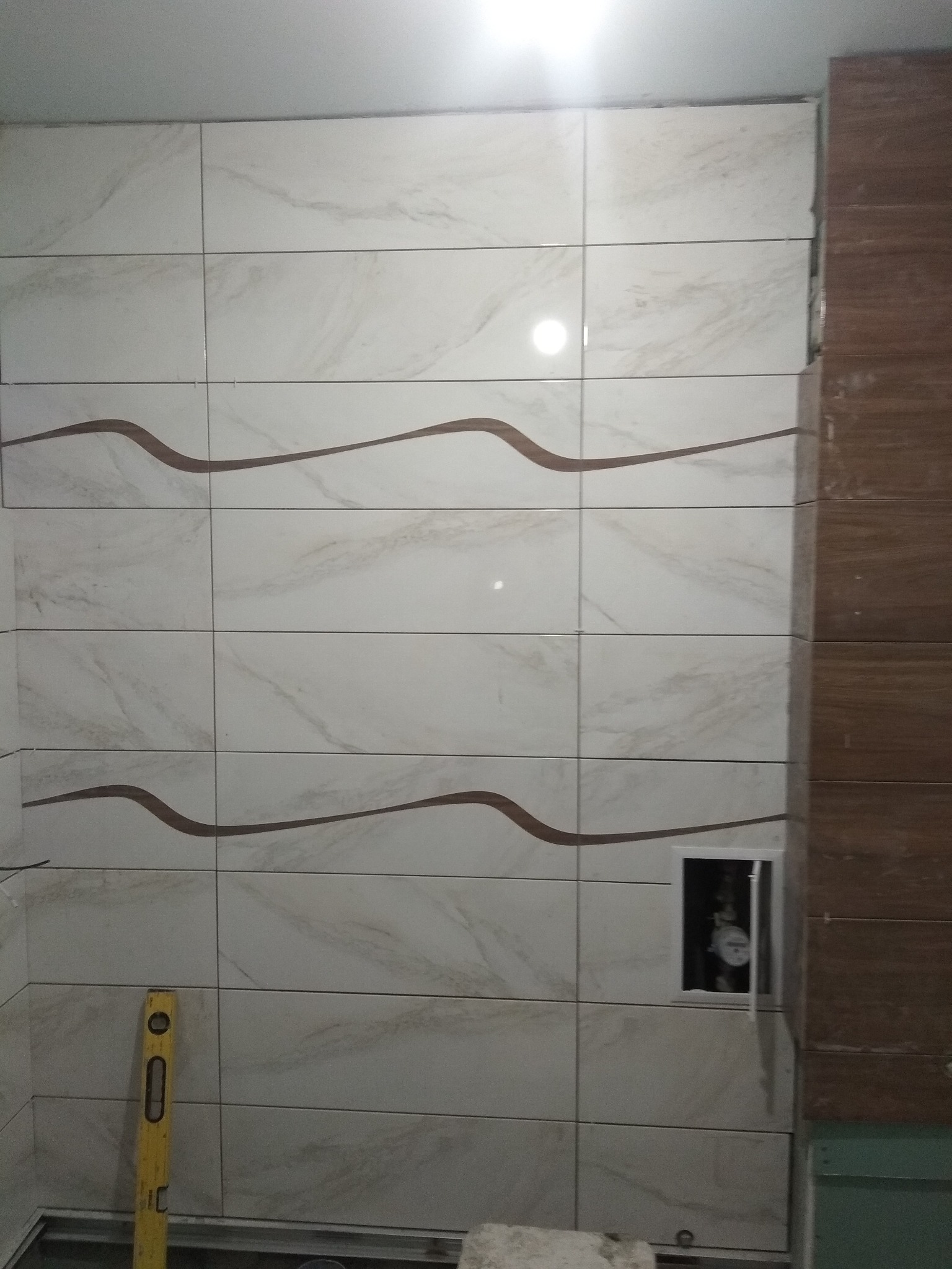 Ściana łazienki wyłożona białymi płytkami z brązowym motywem fal, widoczny pionowy poziomica i otwór rewizyjny z zaworem w środku.