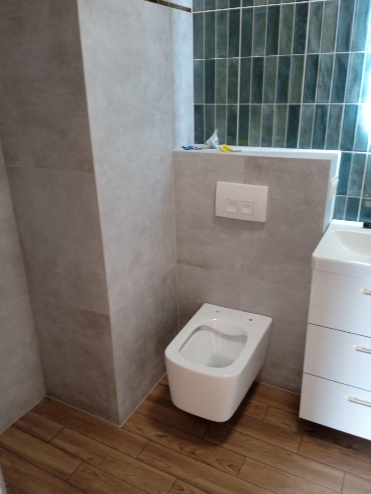 Nowoczesna łazienka z podwieszaną toaletą, szarymi płytkami ściennymi, pionowymi zielonymi płytkami w tle i drewnopodobną podłogą z paneli.