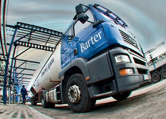 Ciężarówka cysterna z napisem 'Dostawy gazu' i logo 'Barter' na tle instalacji przemysłowej i pochmurnego nieba, widziana z dołu.